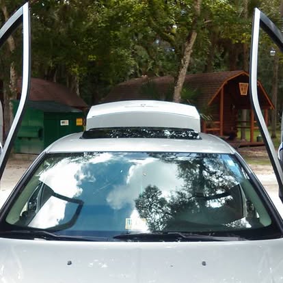 White car with roof rack parked near green and red buildings among trees