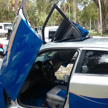 Blue and white car with open scissor doors in a parking lot with trees
