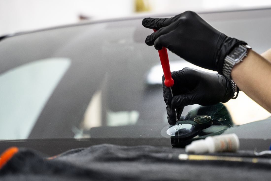 Technician in black gloves applying red resin to windshield chip repair kit on car